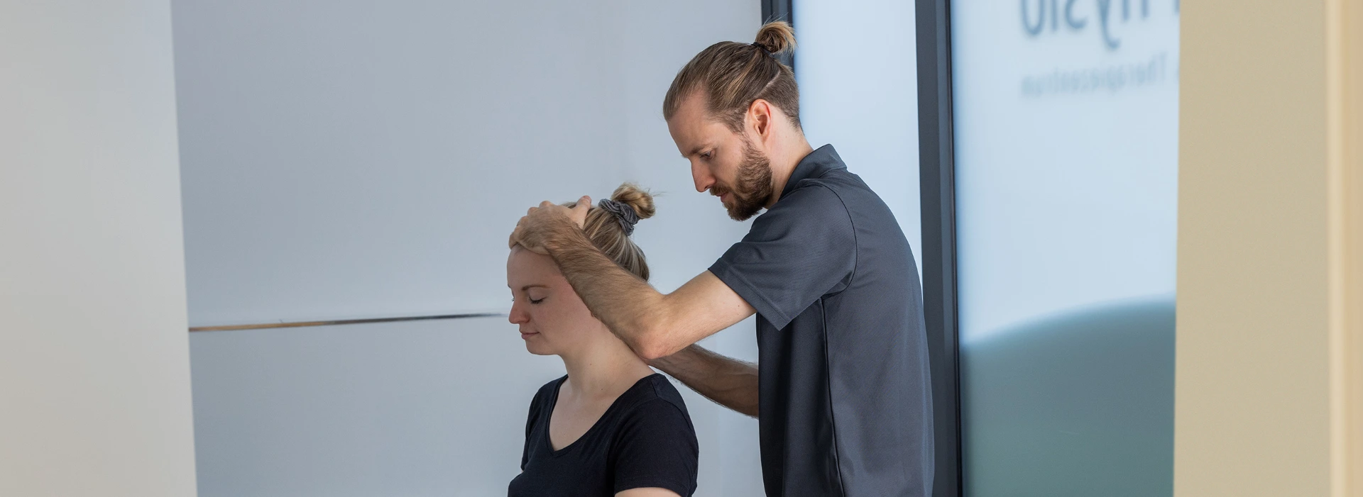 Ein Therapeut führt eine sanfte manuelle Therapie am Kopf einer sitzenden Patientin durch.