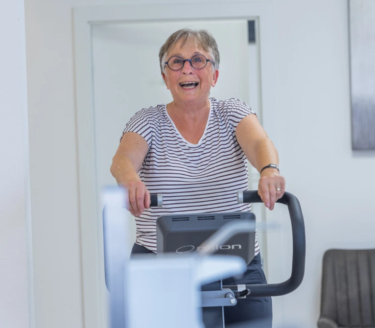 Eine ältere Frau mit kurzen grauen Haaren und Brille trainiert lachend auf einem Heimtrainer in einem hellen Raum. Sie trägt ein gestreiftes T-Shirt und wirkt glücklich und energiegeladen.