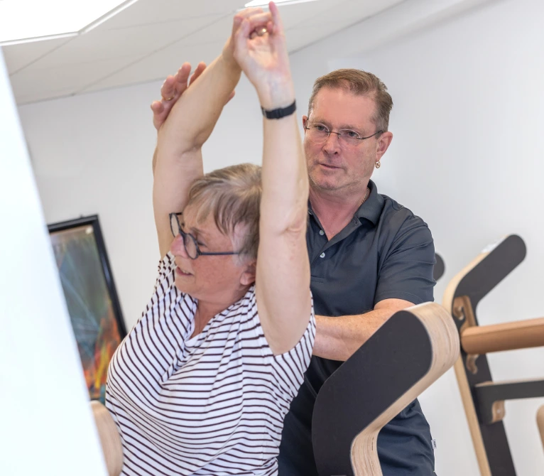 Eine ältere Frau mit gestreiftem T-Shirt streckt ihre Arme nach oben, während ein Mann in dunklem Poloshirt sie bei einer Übung unterstützt. Beide befinden sich in einem hellen Raum mit Fitnessgeräten im Hintergrund.