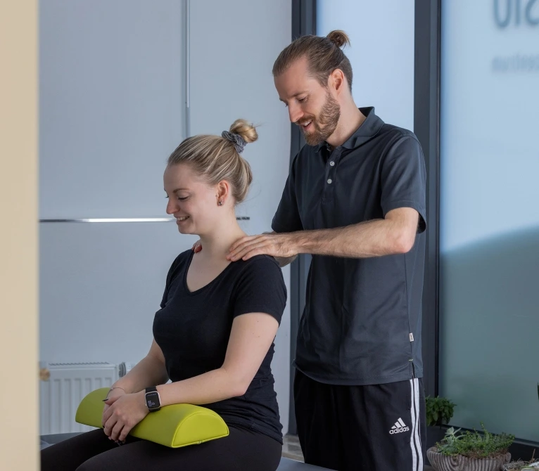 Ein Physiotherapeut führt eine entspannende Schulterbehandlung bei einer sitzenden Patientin durch.