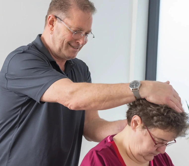 Ein Physiotherapeut führt eine Nacken- und Kopfbehandlung bei einer sitzenden Patientin durch.